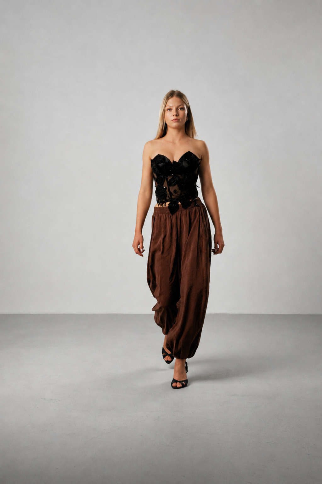 Model trägt ein schwarzes Spitzenkorsett mit floraler Stickerei und brauner Ballonhose