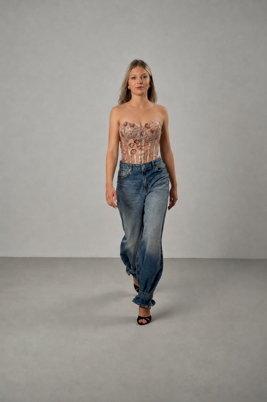 Model trägt ein Spitzenkorsett mit floraler Stickerei in Champagner, kombiniert mit einer blauen Jeans und schwarzen Sandaletten.